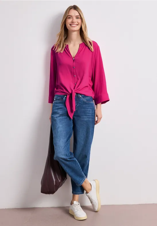 Vrouw in fuchsia blouse met knoop voor, losse blauwe jeans en witte sneakers tegen een lichte muur.