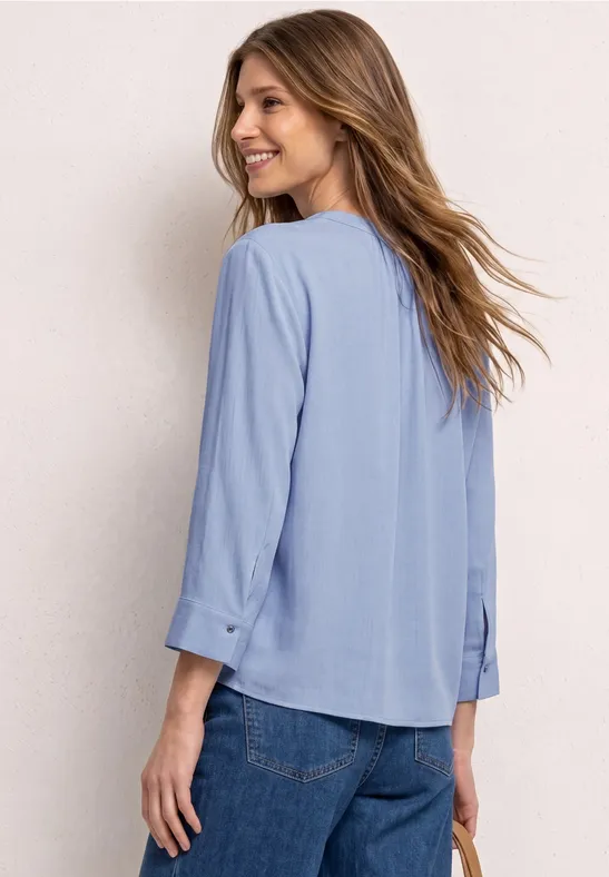 Femme de dos en chemisier bleu clair à manches 3/4 et jean, fond lumineux.