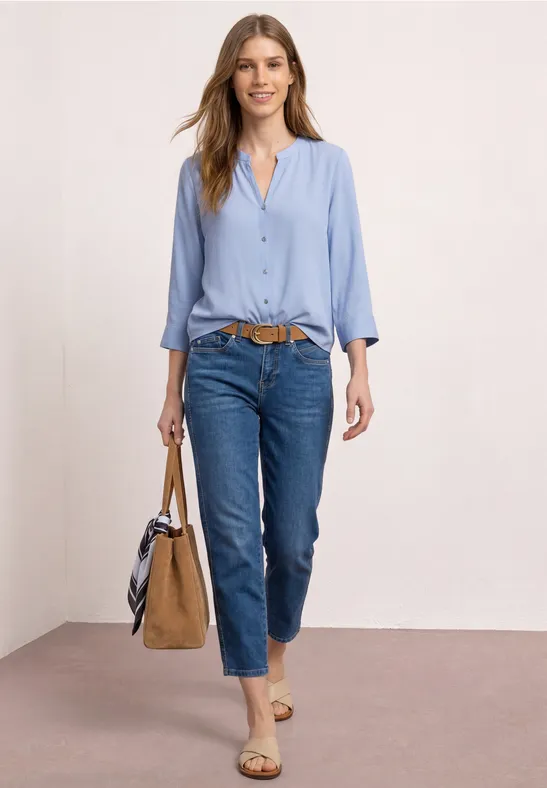 Femme en chemise bleu clair, jean bleu foncé, ceinture marron, sandales et sac à main.