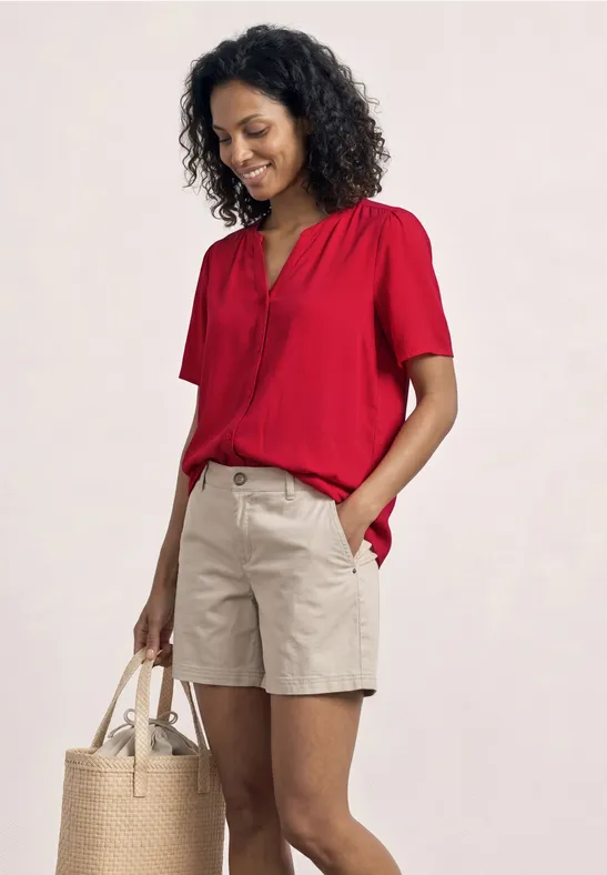 Eine Person trägt ein rotes Kurzarmshirt, beige Shorts und hält eine Strohtasche.