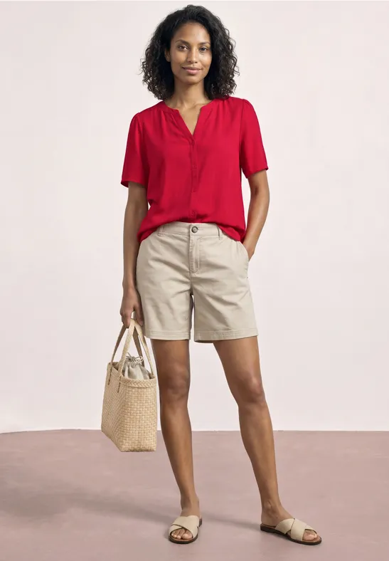 Eine Person in roter Bluse, beigen Shorts und Sandalen trägt eine Strohtasche vor hellem Hintergrund.