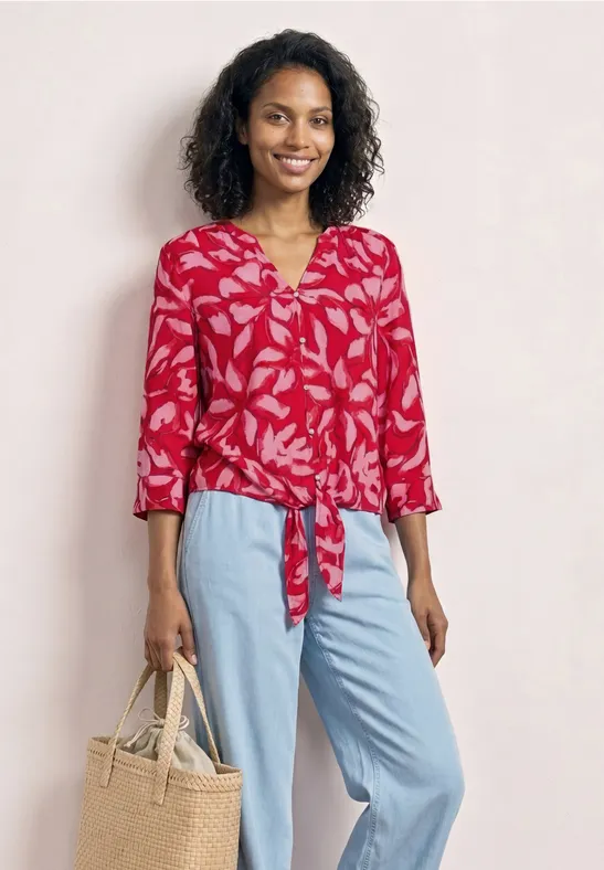 Vrouw draagt rode gebloemde blouse met knoop, lichtblauwe broek en beige tas.