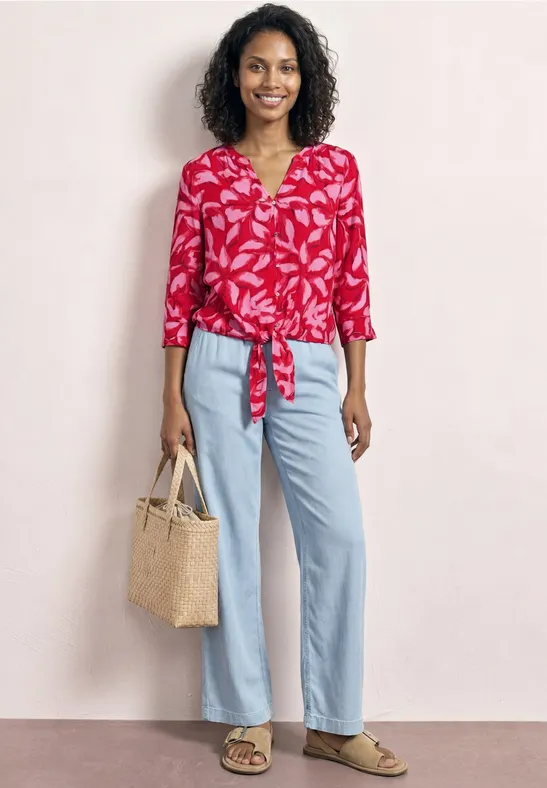 Vrouw in rood gebloemde blouse geknoopt aan de voorkant, wijde lichtblauwe jeans, sandalen en strozak.