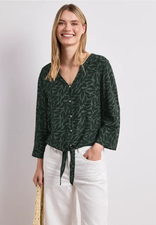 Vrouw in donkergroene geprinte blouse met knoopdetail en witte broek.
