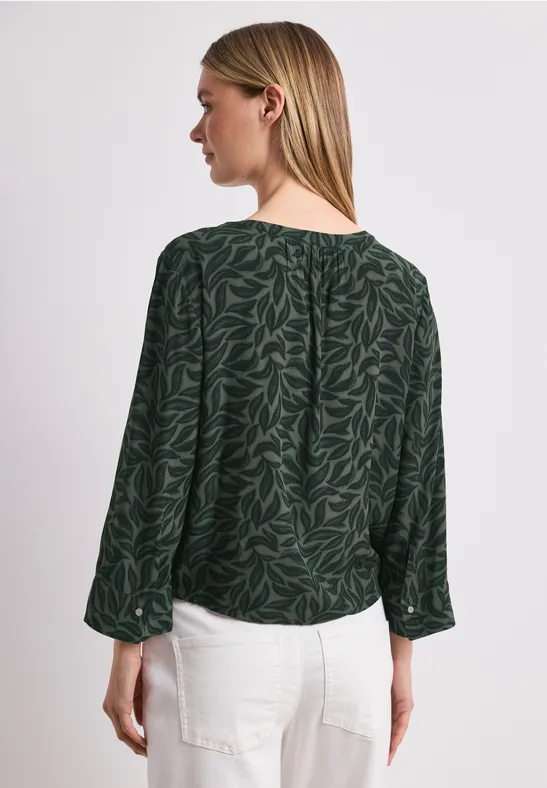 Vrouw van achteren, gekleed in een donkergroene blouse met bladpatroon en een witte broek.