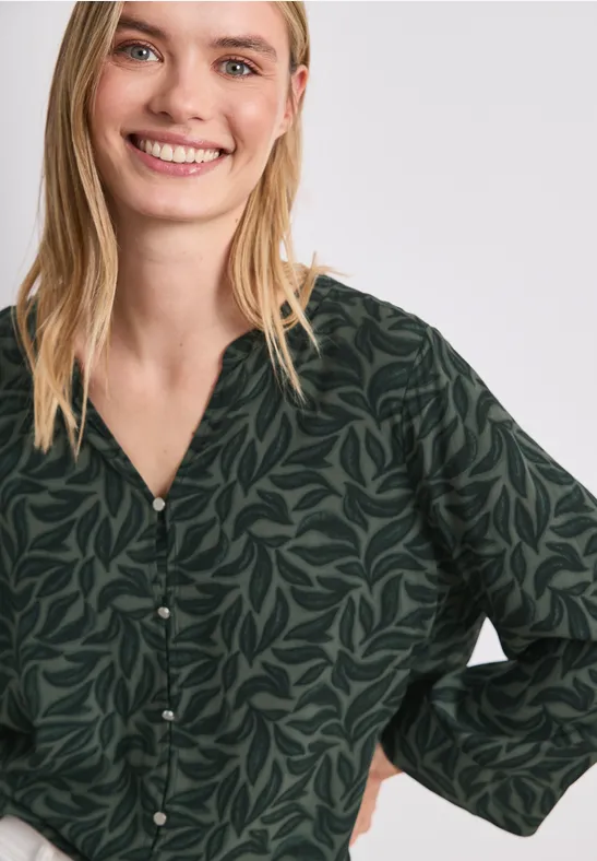 Een glimlachende vrouw draagt een donkergroene blouse met bladmotief.