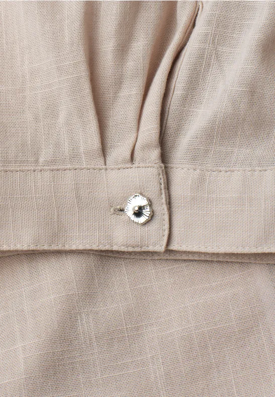 Detail eines beigen Leinenkleides mit Zierlasche und funkelndem Strassknopf.