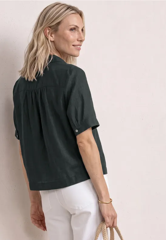 Person steht seitlich mit schwarzem Hemd und weißer Hose, hält Clutch, Blick abgewendet.