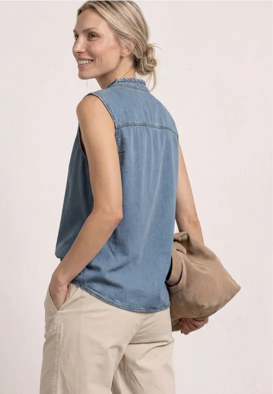 Frau trägt ärmellose Jeansbluse und helle Hose, hält Tasche in der Hand.