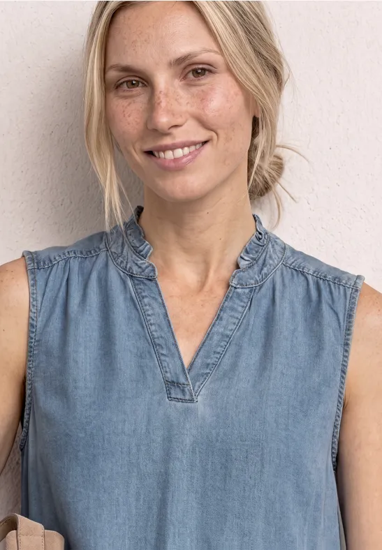 Dame in ärmelloser Jeansbluse mit Rüschenkragen vor hellem Hintergrund.