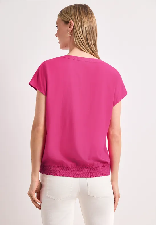 Frau, Rückansicht, pinkes Kurzarmshirt mit Gummizug am Saum, weiße Hose, weißer Hintergrund.
