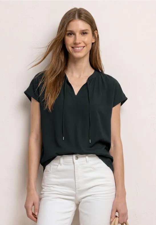 Frau mit schwarzer Kurzarmbluse und hellen High-Waist-Jeans vor hellem Hintergrund.