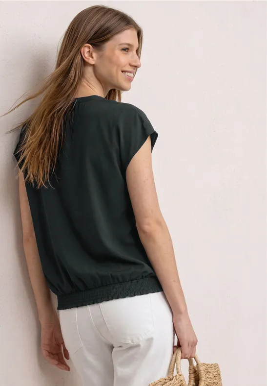 Frau mit schulterlangem Haar, schwarzem Shirt, weißer Hose und Strohtasche steht vor heller Wand.
