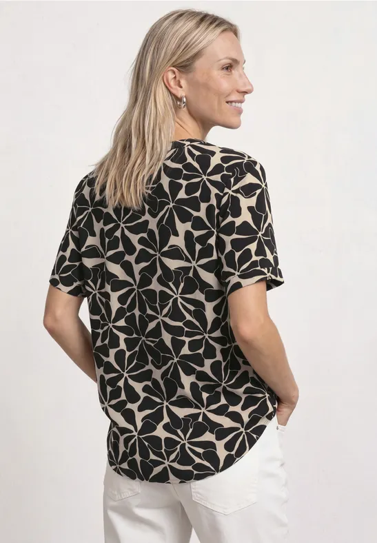 Femme de profil, regard par-dessus l'épaule, chemisier noir-beige à motif et pantalon blanc.