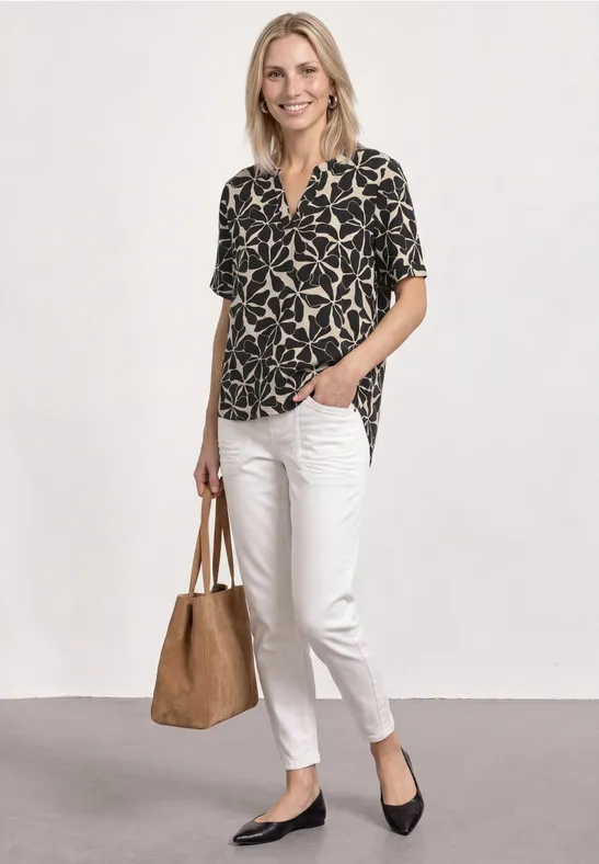 Femme en chemisier à motif, pantalon blanc, ballerines noires et sac cabas marron.