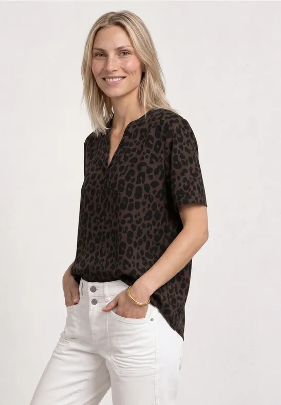 Frau mit kurzärmliger Bluse im Leopardenmuster und weißer Jeans, Hände in den Taschen.
