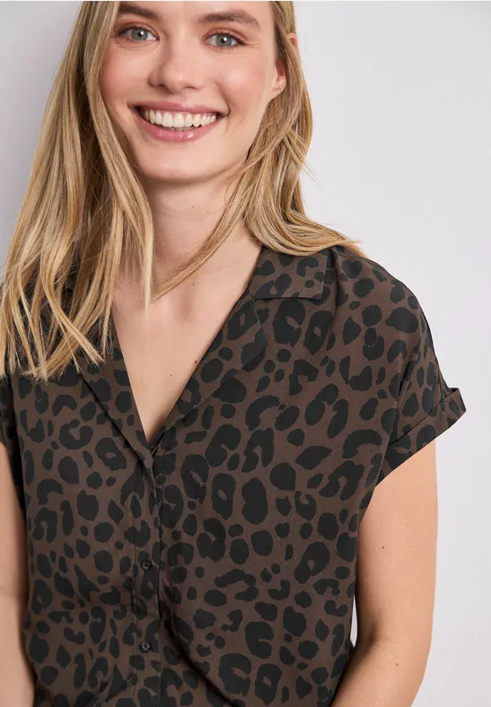 Jonge vrouw met blond haar draagt een donkerbruine blouse met zwarte luipaardprint en lacht.