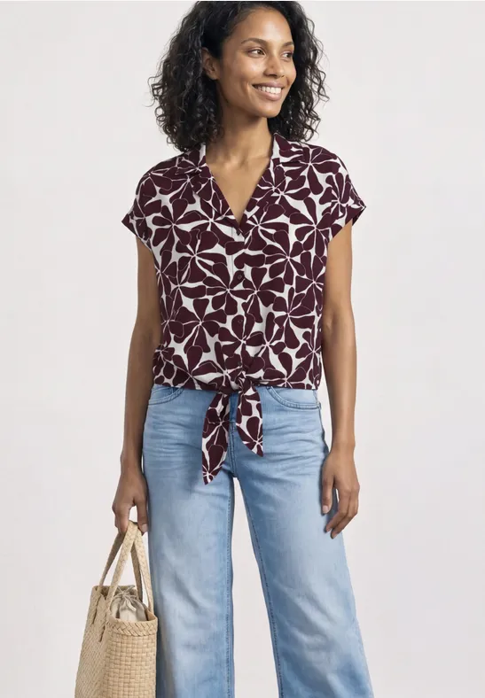 Vrouw in bordeauxblauw gebloemd kort shirt met strik, lichtblauwe jeans en beige tas.