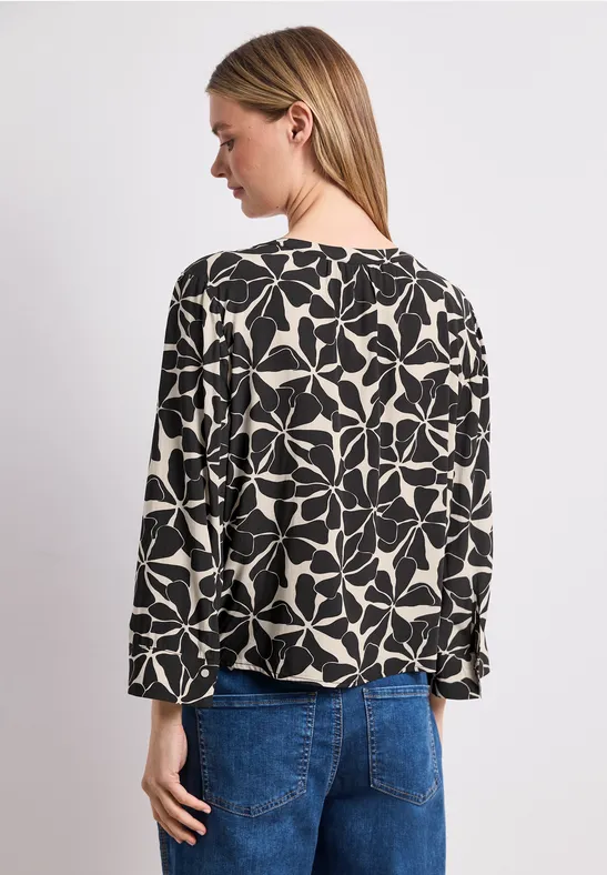 Vrouw van achteren in zwart-wit gebloemde blouse en blauwe spijkerbroek, hoofd licht gedraaid.