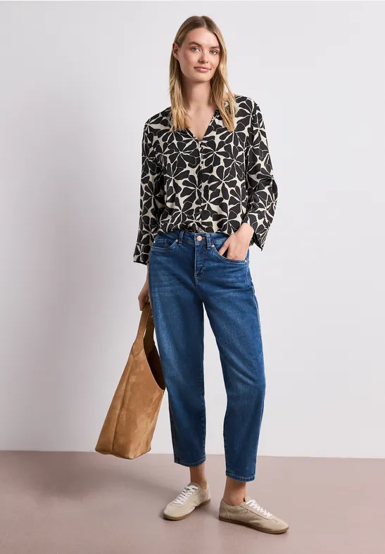 Vrouw met motiefblouse, blauwe jeans en beige schoenen houdt grote bruine tas voor een witte achtergrond.