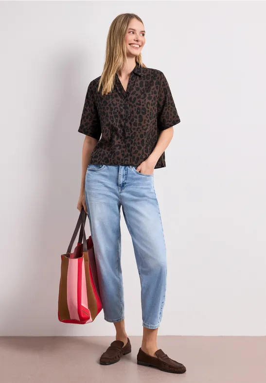 Frau in gemusterter Bluse, hellblauen Jeans, braunen Mokassins und bunter Tragetasche.