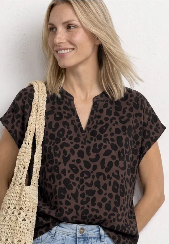 Frau im Leopardenmuster-Top, trägt Jeans und gehäkelte Tasche, lockere Haltung vor weißer Wand.