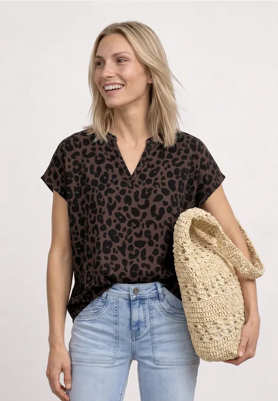 Frau trägt braune Leo-Bluse, helle Jeans und hält eine geflochtene Strohtasche vor hellem Hintergrund.