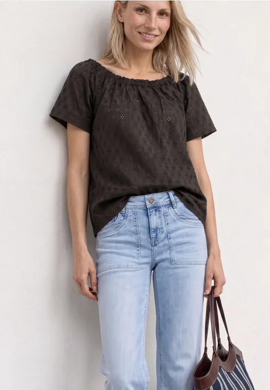 Frau mit schwarzer Bluse, hellblauen Jeans und brauner Handtasche vor weißem Hintergrund.