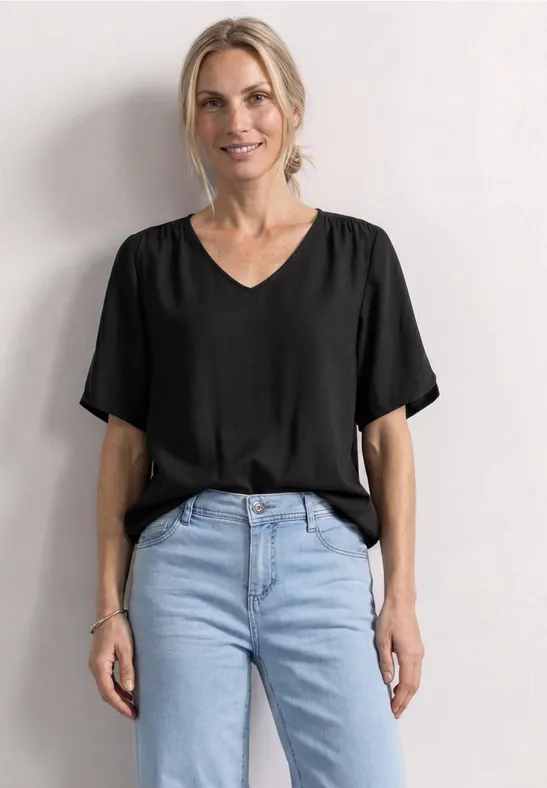 Frau in schwarzem V-Ausschnitt-Shirt und hellblauer Jeans vor weißem Hintergrund.