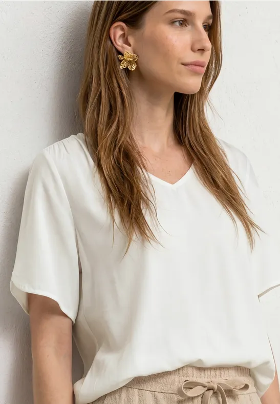 Frau in weißer V-Ausschnitt-Bluse und beigen Hosen, mit goldenen Blumen-Ohrringen und langem braunem Haar.