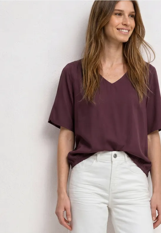 Frau in bordeauxfarbenem V-Shirt und weißer High-Waist-Hose vor hellem Hintergrund.