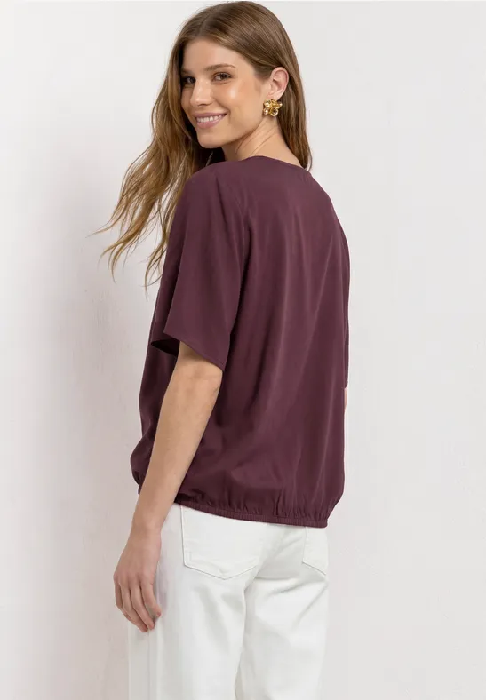 Seitlich abgelichtete Person trägt weinrotes T-Shirt und weiße Hose vor weißem Hintergrund.