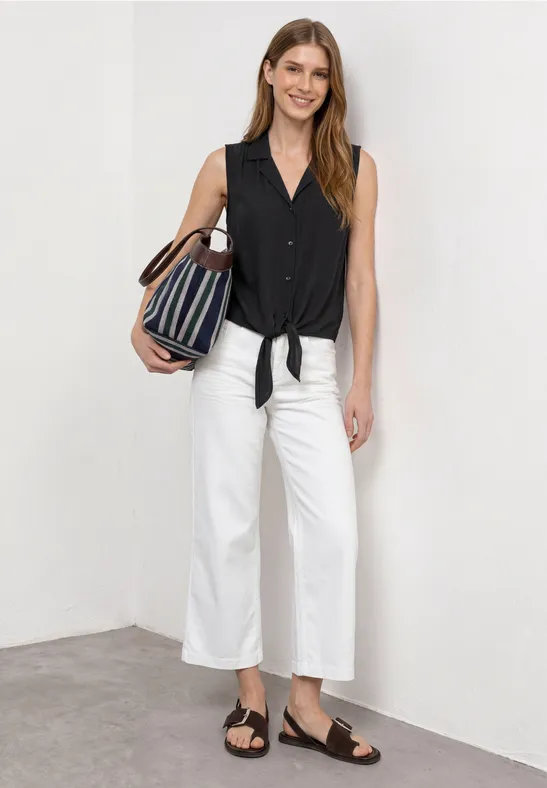Frau trägt schwarzes ärmelloses Top mit Knotendetail, weiße Culottes, schwarze Sandalen und gestreifte Tasche.