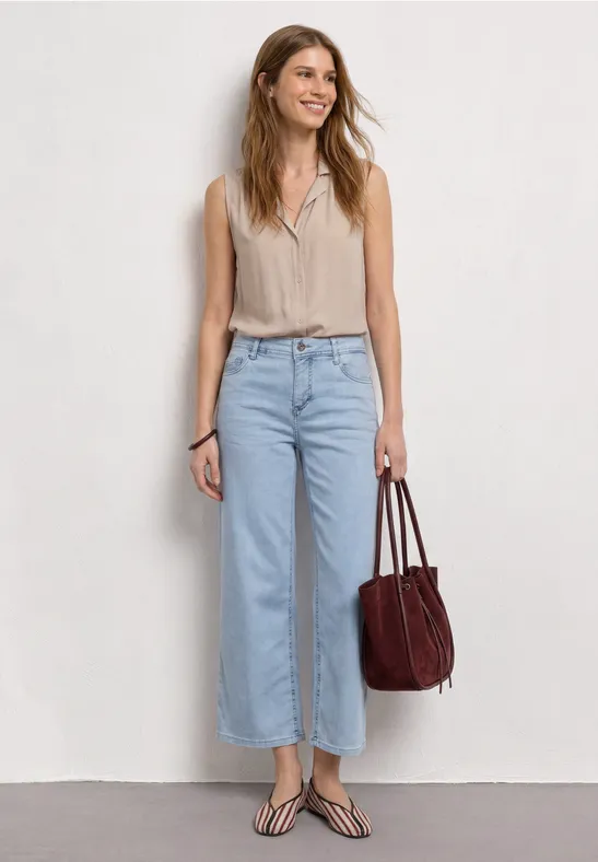 Frau trägt ärmelloses Beige-Oberteil, hellblaue, weite Jeans, Zehentrenner-Sandalen und eine braune Tasche.