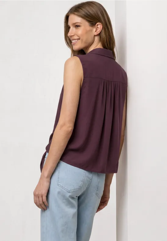 Eine Frau von hinten in ärmelloser lila Bluse und hellen Jeans vor weißem Hintergrund.