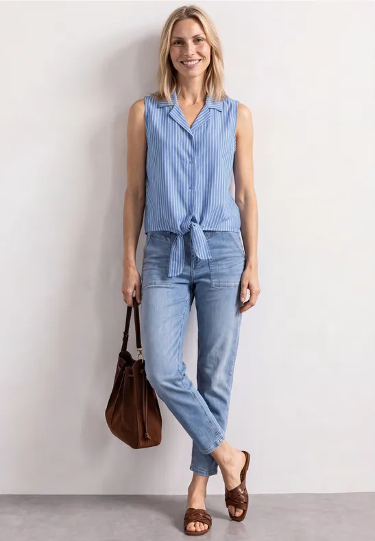 Vrouw in lichtblauwe denim, mouwloos hemd knoopt in de taille, ruime jeans, bruine leren tas en gevlochten sandalen.