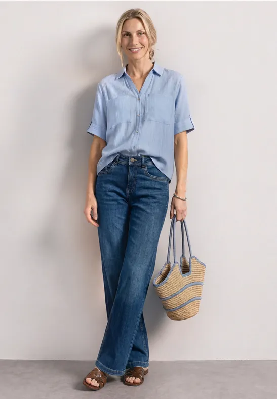 Vrouw in lichtblauw overhemd, wijde jeans en sandalen met een gestreepte strozak tegen een neutrale achtergrond.