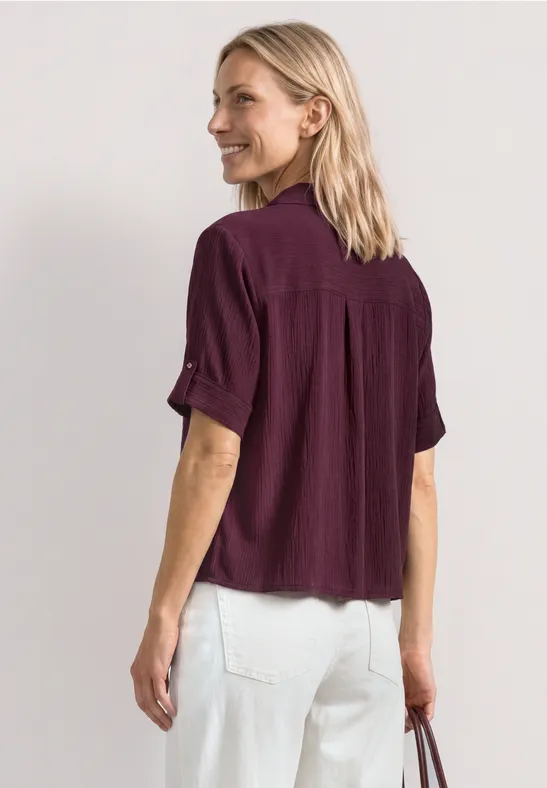 Vrouw van achteren met een paarse blouse, witte spijkerbroek en een tas in haar linkerhand.