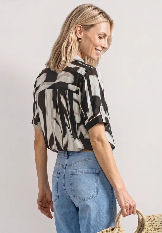 Frau mit Zebramuster-Bluse, hellen Jeans und Strohtasche, Blick abgewandt.