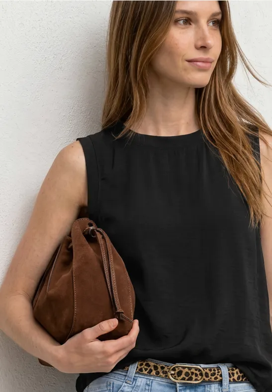 Schlichtes Outfit: Frau in schwarzem Top, brauner Tasche und Gürtel lehnt an weißer Wand.