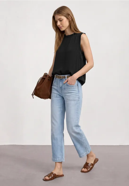 Frau in schwarzem Top, blauen Jeans und braunen Sandalen mit passender Tasche vor hellem Hintergrund.