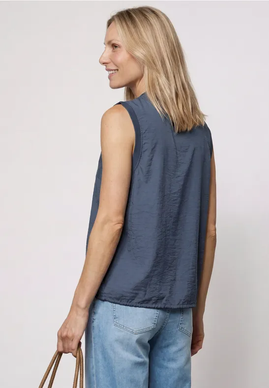 Frau mit blondem Haar von hinten, trägt ein ärmelloses dunkelblaues Oberteil, Jeans und hält eine kleine Tasche.