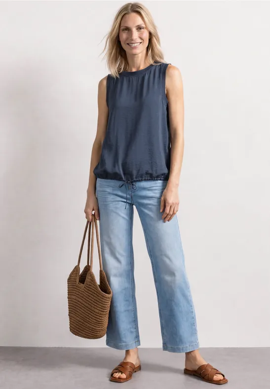 Frau trägt ärmellose Bluse, hellblaue Jeans, braune Sandalen und Korbtasche.