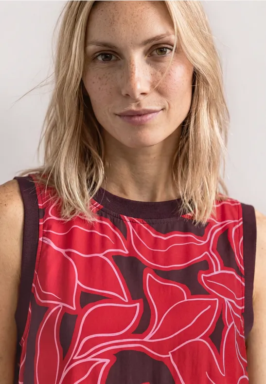 Blonde vrouw draagt rood mouwloos topje met abstract, bloemig patroon voor een neutrale achtergrond.