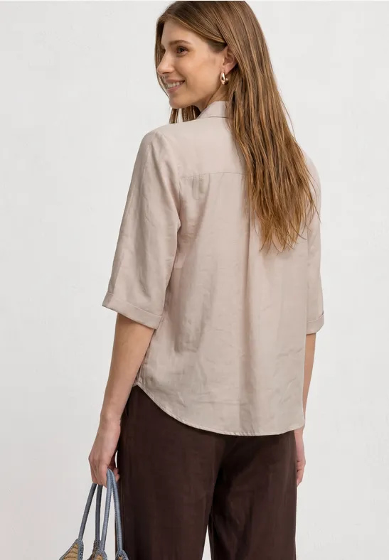 Frau von hinten in einer beigen Hemdbluse und dunklen Hosen, Handtasche in der Hand.