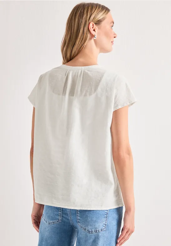 Vrouw van achteren in een wit kort mouwtoppje en een blauwe jeans voor een lichte achtergrond.
