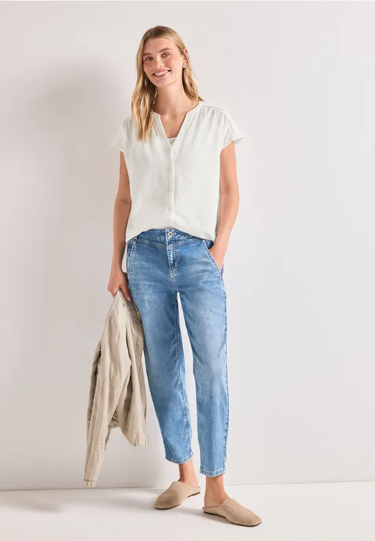 Een vrouw in een wit overhemd, blauwe jeans en loafers houdt een lichte jas in haar hand.