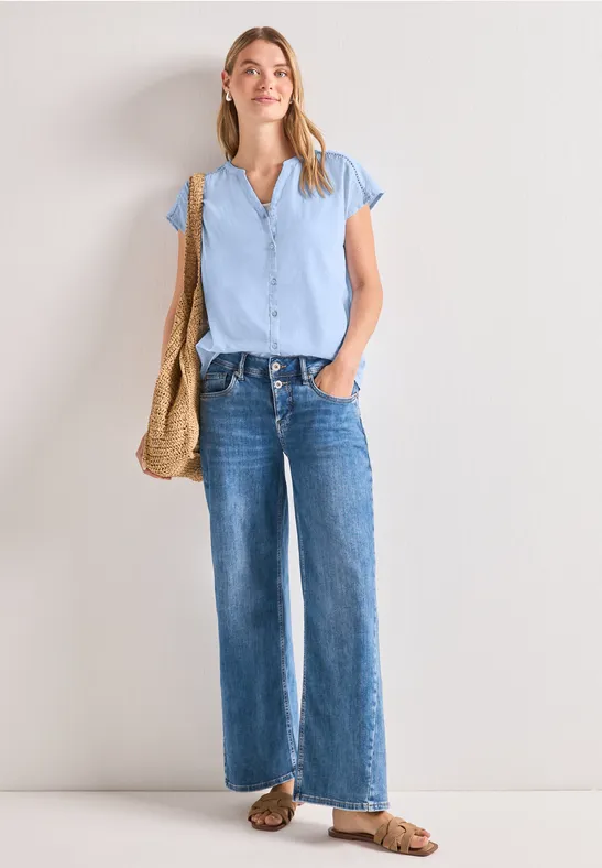 Een vrouw draagt een lichtblauw T-shirt, wijde spijkerbroek, sandalen en een rieten tas voor een witte muur.