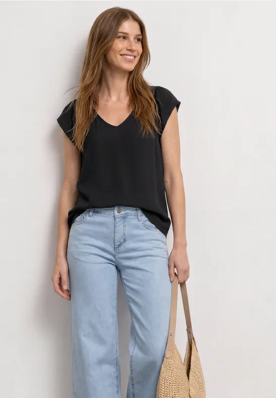 Femme en haut noir à col V, jean bleu clair, tenant un sac en paille.