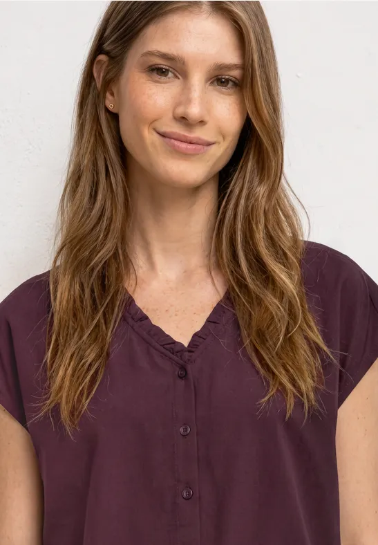 Vrouw met lange, golvende haren draagt een lichtbruine blouse met korte mouwen voor een witte achtergrond.
